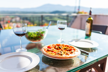 Fresh handmade pizza on glass table on terrace outside in Italy with tomato sauce vegetables and two empty white plates and glasses of red wine green salad