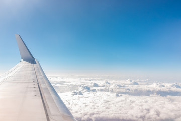 White blue airplane in sky with view from window high angle during sunny day with plane wing and clouds covering horizon