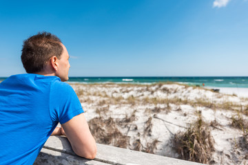 Fototapeta na wymiar Destin Miramar beach city town village in Florida panhandle gulf of mexico ocean with back of young man closeup in blue shirt leaning on fence railing by sand dunes