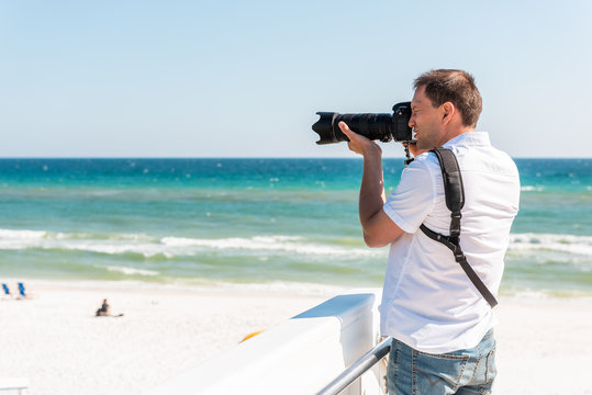 Young photographer man taking picture on beach during sunny day in Seaside, Florida panhandle town village with ocean by steps