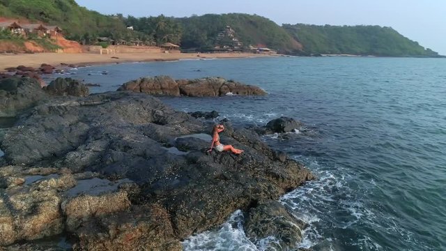 Aerial view. Flying around a young cute woman sitting on the edge of a stone reef that is washed by the waves she looks at the sunset over the sea, covering her eyes with her hand.