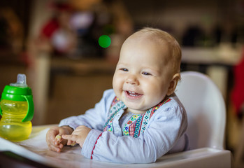 Cute baby girl sitting in high chair and eating piece of bread