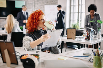 Young redhead businesswoman drinking coffee while working in the office.