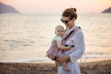 Cheerful Caucasian woman with little daughter in baby carrier on beach at sunset