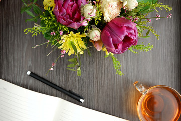 Bright bouquet of flowers with notepad and tea on a wooden table, top view