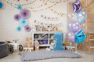 Birthday blue and purple decorations with gifts, toys, garlands and figure for little baby party on a white bricks background.