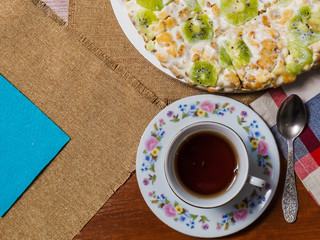 A cake made from biscuits, decorated with kiwi slices, lies in a white plate next to a saucer and a cup of black tea on checkered napkins.