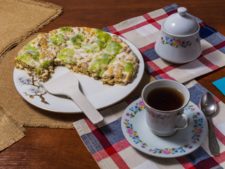 A cake made from biscuits, decorated with kiwi slices, lies in a white plate next to a saucer and a cup of black tea on checkered napkins.