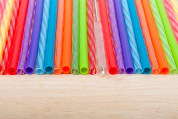 Row of colorful plastic and silicone reusable straws with cleaning brush in the middle laying on light wood table, viewed from above in front. Ecologically friendly.