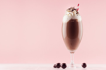 Sweet chocolate milkshake in exquisite wineglass decorated candy balls, whipped cream, chocolate chips, straw on light pastel pink background.