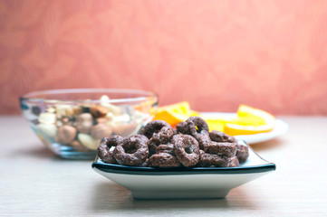 Crunchy Chocolate Cereals Round Oats in a Dish, Glass Bowl of Nuts and Orange Slices in a White Plate on Wooden Table. Pink Textured Background. Healthy Organic Snack, Food, Breakfast Concept.