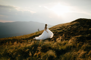 Fototapeta premium Stylish bride standing on beautiful landscape of mountains on sunset