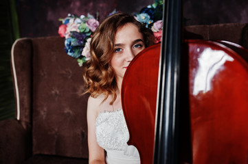 Pretty young gilrl musician in white dress with double bass sitting on brown vintage sofa.