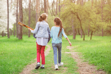 two happy free young girls with guitar walking singing song along spring green park pathway