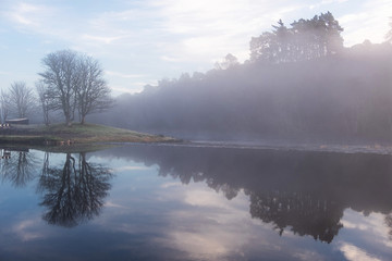 Fototapeta premium A lonely island reflected in the glassy smooth surface of a lake shrouded in morning fog