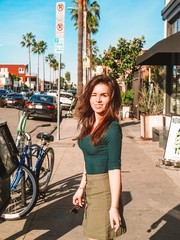 A girl with long hair and a skirt stands on a Los Angeles street with simple houses and a Bicycle