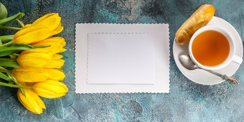 Horizontal banner. White blank card, yellow tulips, cup of tea on blue concrete background. Top view