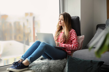 Beautiful young woman with laptop sitting on window sill