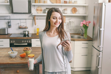 Beautiful woman drinking milk and using mobile phone in kitchen at home