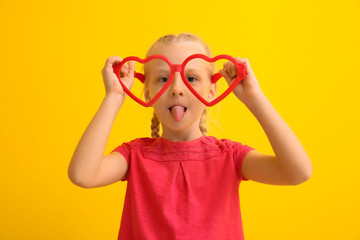 Funny little girl with big heart-shaped glasses on color background. April fools' day celebration
