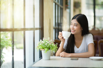 A beautiful woman who is sitting drinking coffee near the window with warm sunlight passing through in the morning.