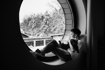 The boy on the window in his pajamas. Holds a black cat in his hands. Beautiful view from the round window. Home comfort. Interior. The layout of the house. Black and white photo