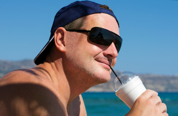 Smiling unshaven male in sunglasses drinking a cocktail on the sea coast