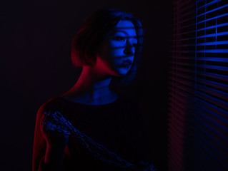 A beautiful girl with a chain in her hands stands near the window with blinds in the light of neon lights of blue and red light