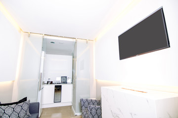 Modern waiting room with white panels and tv in the wall and technological marble fireplace. There is a small kitchen at the end of the hall.