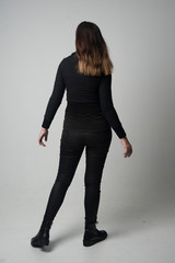 full length portrait of a brunette girl wearing  modern black jacket and pants, standing pose with back to the camera on grey studio background.