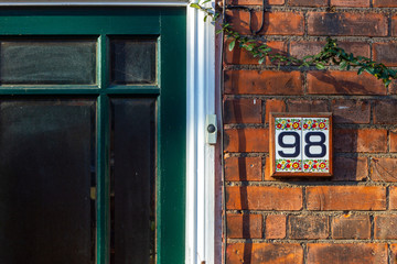 House number 98 on bright painted tiles next to a door bell and a green house door. The ninety eight on tiles on a brick wall