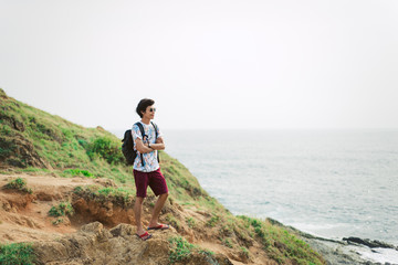 Fototapeta premium Young man traveler with sunglasses posing in a white t-shirt standing on the pier near the sea with a backpack on shoulders, success, freedom and happiness.
