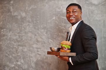 Respectable young african american man in black suit hold tray with double burger against gray wall.