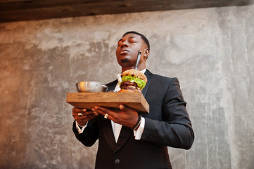 Respectable young african american man in black suit hold tray with double burger against gray...