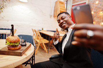 Respectable young african american man in black suit sitting in restaurant and making selfie against tasty double burger and show thumb up.