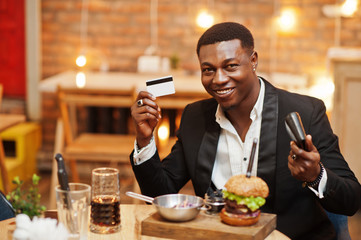 Respectable young african american man in black suit sitting in restaurant hold credit card and wallet with tasty double burger and soda drink.
