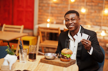 Respectable young african american man in black suit sitting in restaurant hold a lot of credit cards with tasty double burger and soda drink.