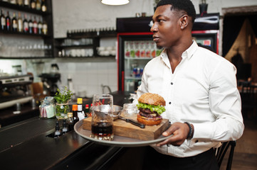 Young african american waiter man hold tray with burger at bar of restaurant.