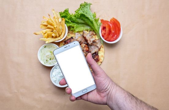 Gyros And Smartphone. Hand Holding A Mobile Phone With Blank White Screen, Gyro Pita Ingredients Background.