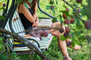 Teen girl reads a book sitting on a garden swing in the summer garden.