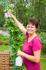 Smiling senior female gardener taking care of plants in summer garden, spraying a plant with pure water from a bottle, vertical
