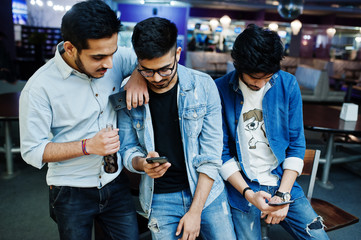 Three stylish asian mans wear on jeans looking at mobile phones and drink soda against table on club.