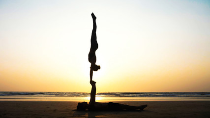 Silhouette of fit sporty couple practicing acrobatic yoga with partner together on the sandy beach....