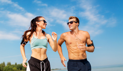 fitness, sport and technology concept - happy couple with earphones and arm bands running along...