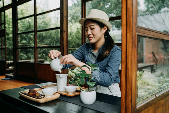 elegant beautiful asian girl doing chadou organic green matcha tea in bowl sitting in japanese wooden house with teien zen garden in background in spring season. woman with hat hands pouring teapot
