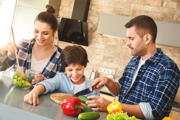 Family at home standing in kitchen together father showing son cutting vegetables while mother mixing salad happy