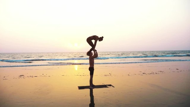 Silhouette of fit sporty couple practicing acrobatic yoga with partner together on the sandy beach. Steadicam shot of female acrobat doing handstand on the hands of his standing male partner. 