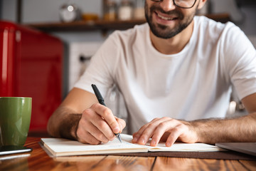 Photo of pleased man writing down notes while using silver laptop on kitchen table