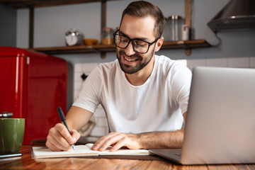 Photo of attractive man writing down notes while using silver laptop on kitchen table