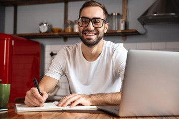 Photo of smiling man writing down notes while using silver laptop on kitchen table